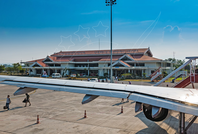Aircraft wing and engine on the apron facing a low red-roofed tropical airport terminal under blue sky