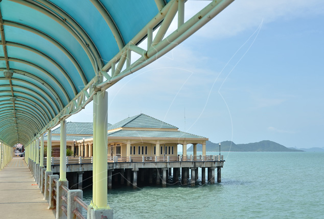 Covered turquoise walkway along a concrete pier leading to a Thai-style terminal building over calm green sea