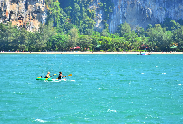Two people paddle a kayak in turquoise sea below limestone karst cliffs and a tree-lined beach with a longtail boat