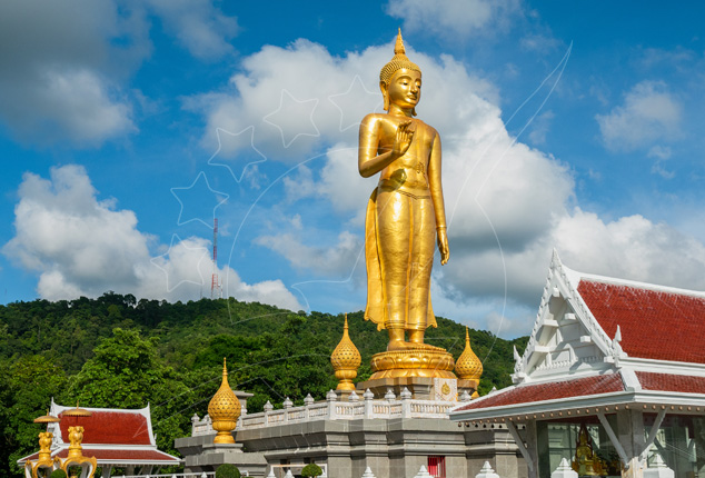 Tall golden standing Buddha statue above a white and red-roofed temple on a forested hill, Hat Yai, Thailand