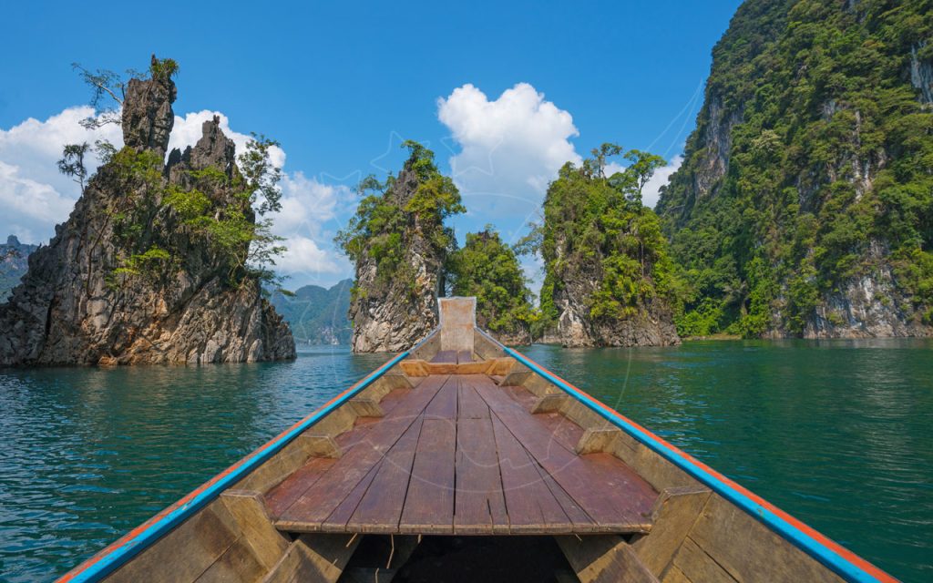 Bow of wooden longtail boat on emerald lake with jungle-covered limestone karst peaks under blue sky