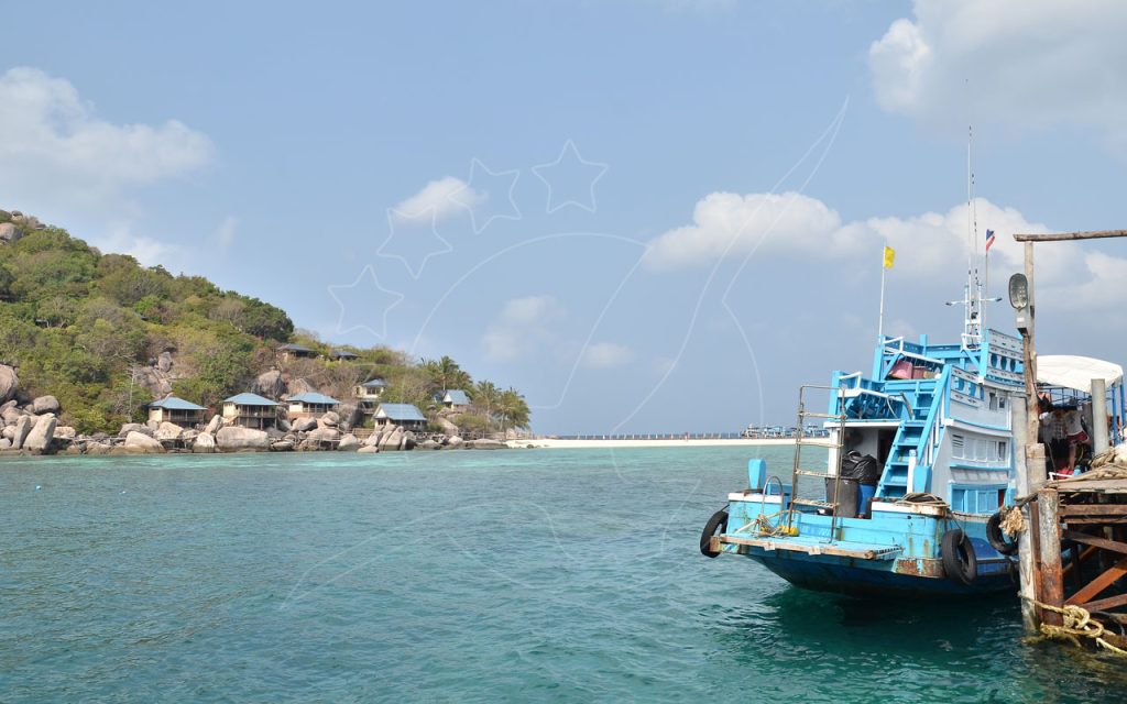 Blue wooden Thai fishing boat moored at a weathered pier beside a turquoise bay with sandbar and hillside bungalows