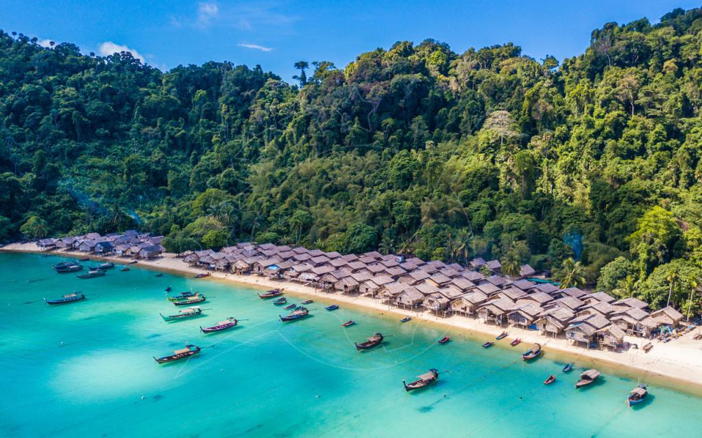 Aerial view of a thatched stilt village on a white-sand beach with longtail boats moored in turquoise water below jungle hills