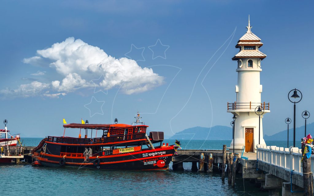 Red and black wooden dive boat moored at a concrete pier beside a white Thai-style lighthouse under blue sky