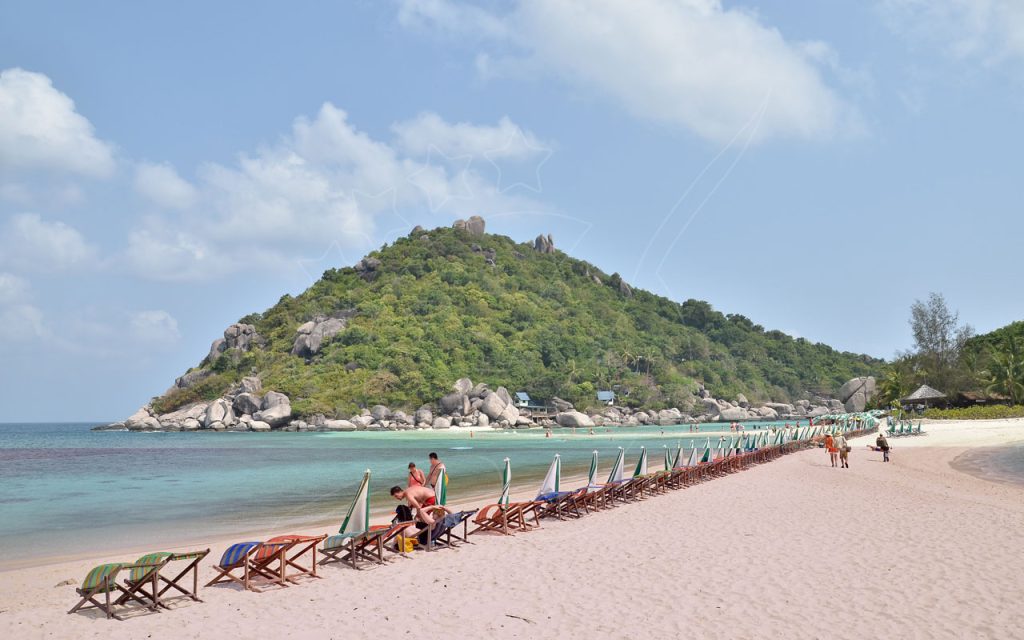เก้าอี้ชายหาดลายทางและร่มที่หุบไว้เรียงยาวบนหาดทรายขาว หันหน้าสู่เกาะหินสีเขียว ประเทศไทย