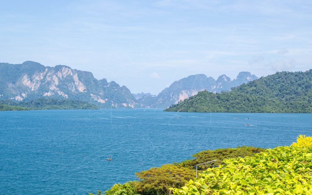 Turquoise reservoir ringed by limestone karst peaks and jungle hills, with small longtail boats and leafy green foreground