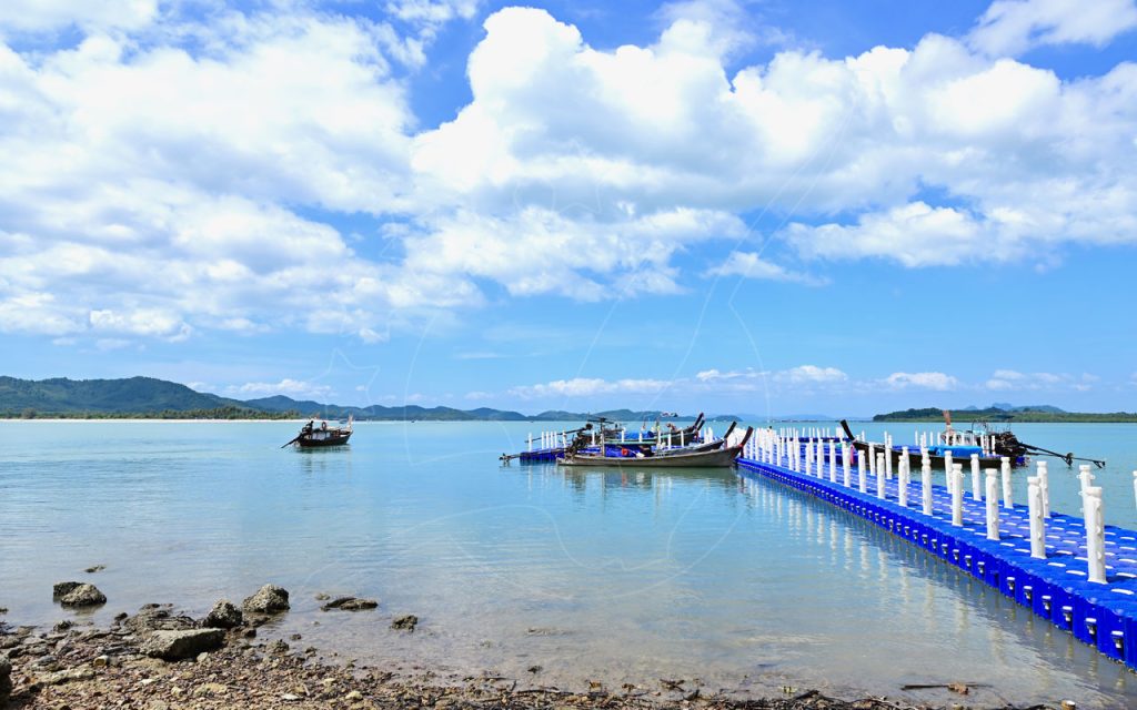 Blauer schwimmender Kunststoffponton mit vertäuten Longtailbooten auf ruhigem türkisem Wasser unter Quellwolken