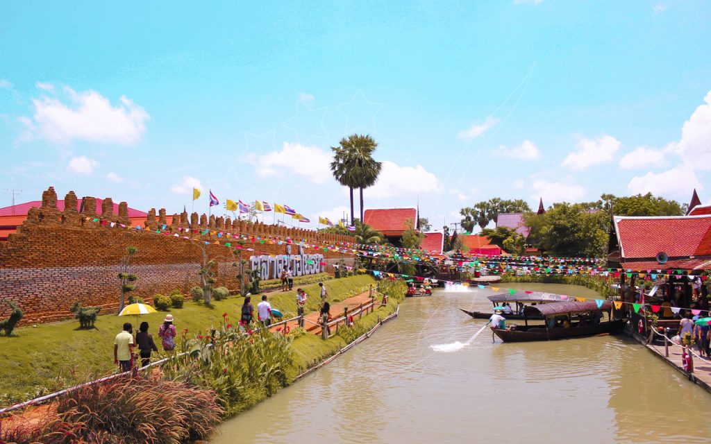 Barche long-tail su un canale accanto a un muro di mattoni e bandierine colorate in un mercato galleggiante thailandese, turisti lungo un sentiero