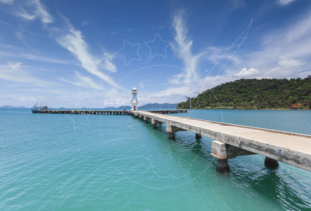 Concrete pier extending over turquoise sea toward a small white lighthouse, with forested island and blue sky