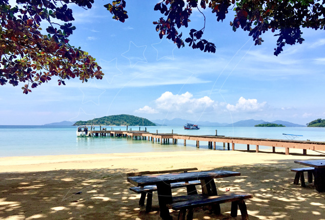 Long wooden pier stretching into turquoise sea past islands, with beach picnic tables shaded by trees