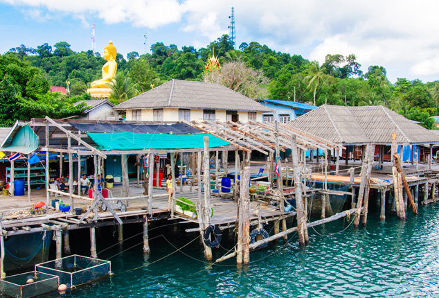 Stilt houses and wooden fishing piers over turquoise water, with a golden Buddha statue on a jungle hill behind