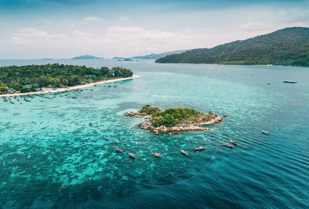 Aerial view of a small rocky islet ringed by longtail boats in turquoise reef water beside a forested beach