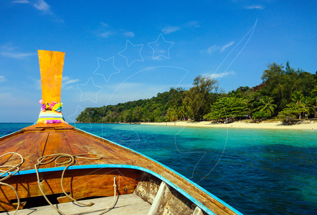 Bow of a wooden longtail boat with flower garlands heading toward a turquoise bay and tree-lined sandy beach