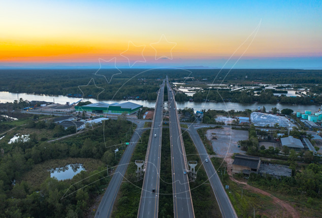 Aerial dusk view of a divided highway bridge crossing a wide river past warehouses and mangrove forest