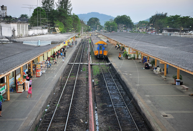 Elevated view of a Thai railway station with two long covered platforms, tracks and a blue-orange diesel railcar.