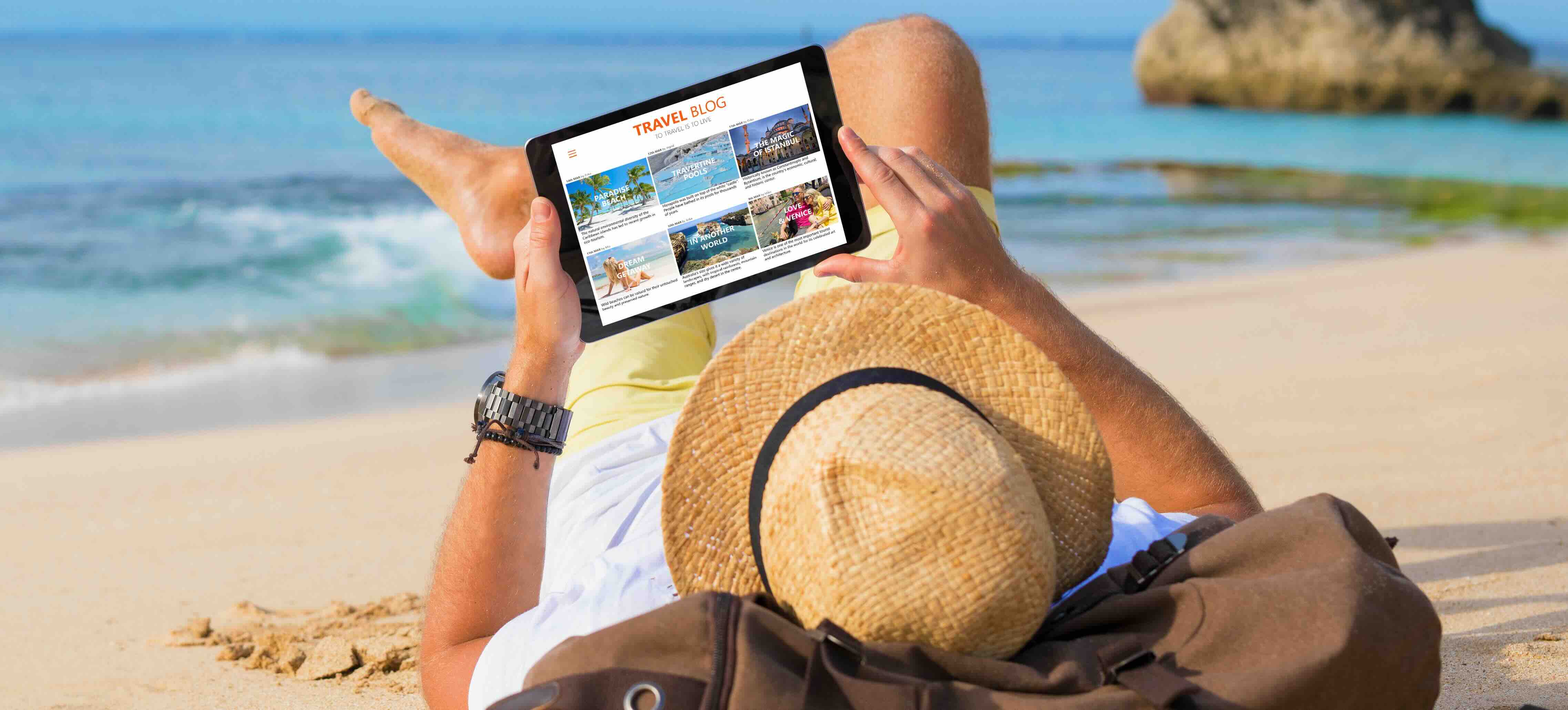 Hombre tumbado en una playa de arena leyendo un blog de viajes en una tableta, con sombrero de paja y camisa amarilla