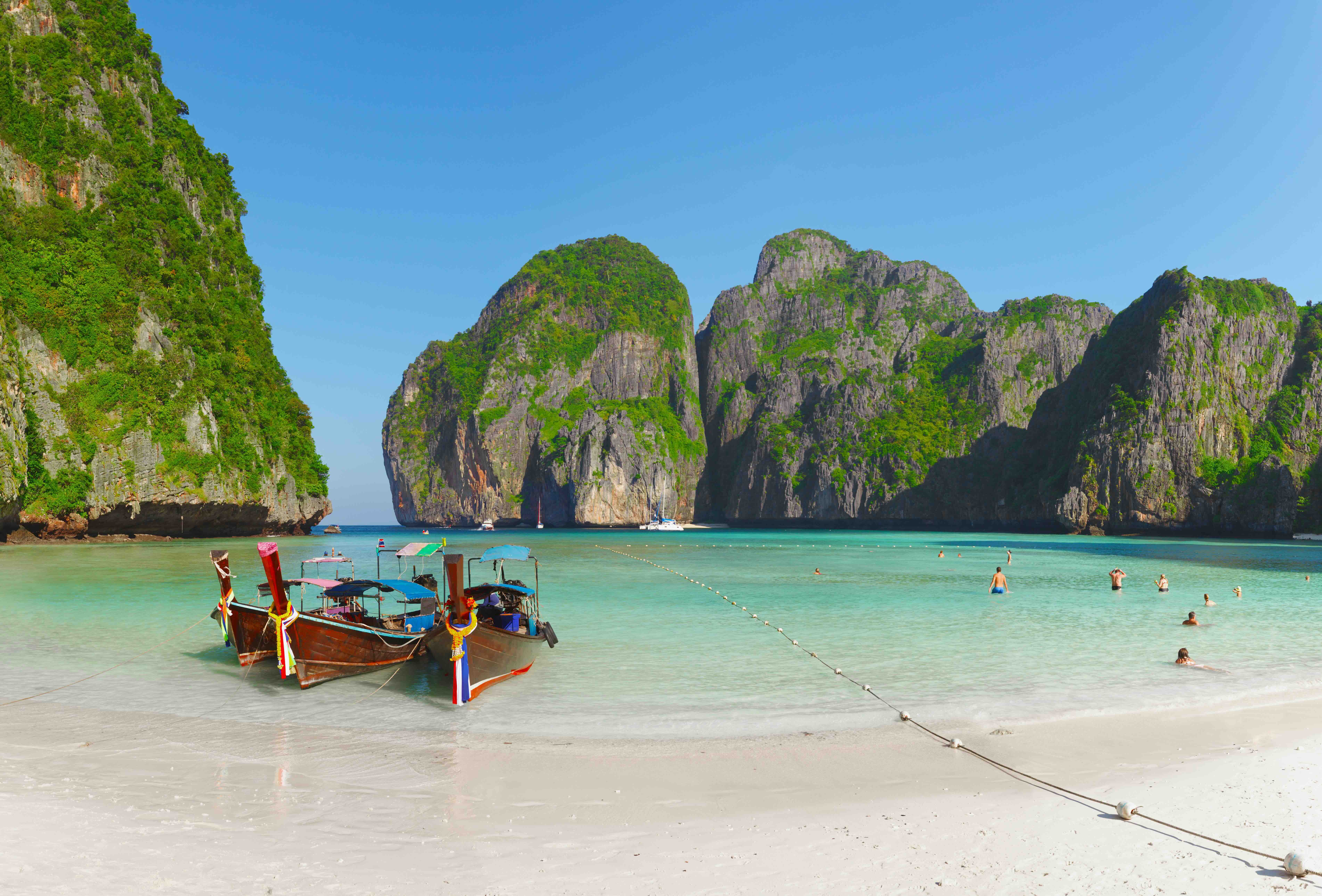 Two wooden longtail boats moored on white sand at a turquoise bay ringed by limestone cliffs, swimmers nearby