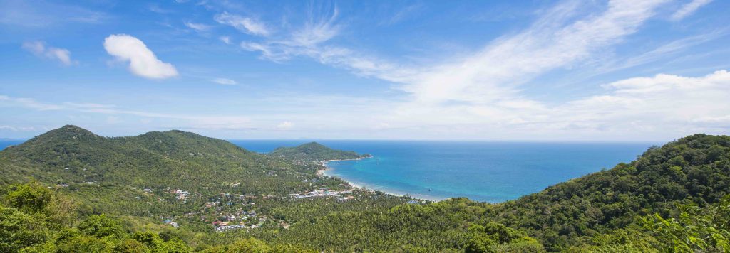 Lush green hills descend to turquoise waters and sandy beach with coastal settlement under blue sky